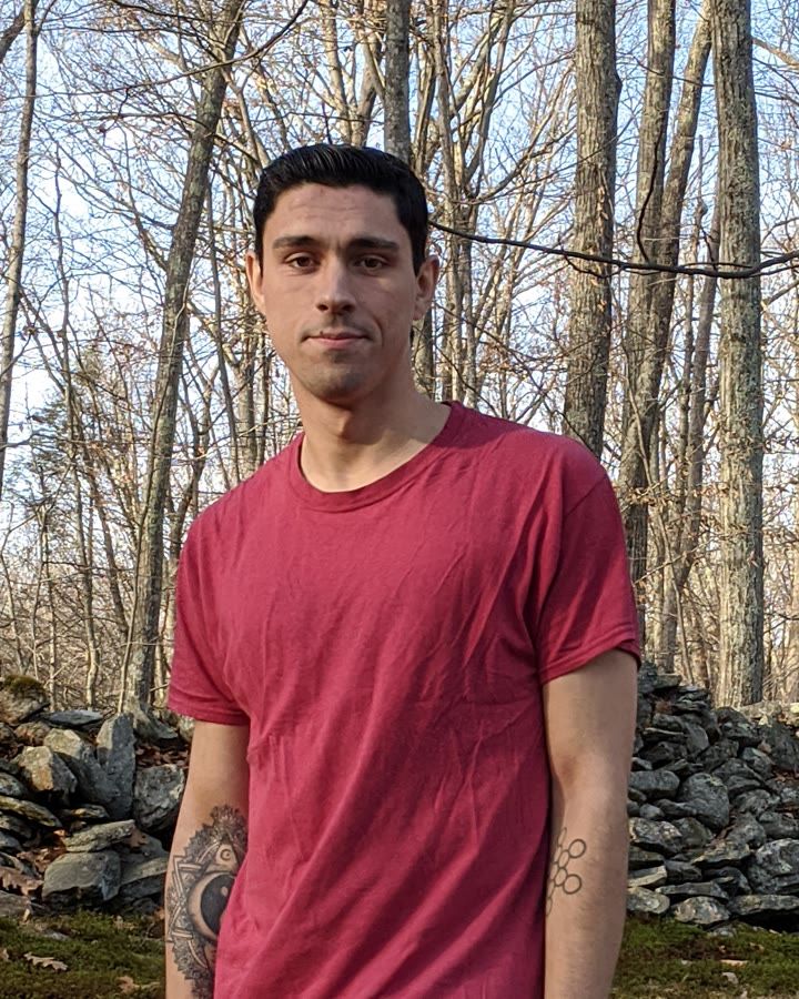 Devin in November 2019, thin and unsmiling, in a red T-shirt in the woods